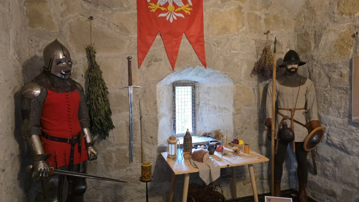 Scena historyczna przedstawiająca grupę rycerzy i postaci średniowiecznych w wnętrzu baszty lub zamku. Widoczne są elementy rekonstrukcji, takie jak zbroje, broń oraz dekoracje nawiązujące do epoki rycerskiej.